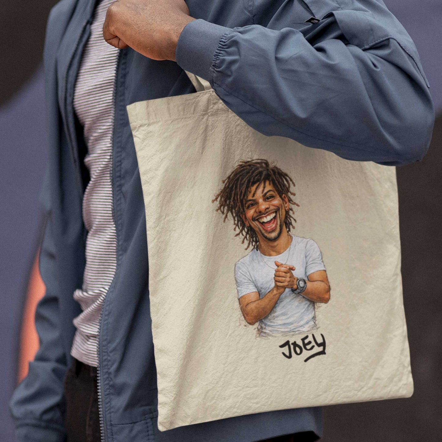 Beige katoenen totebag gedragen over de schouder van een persoon in een blauwe jas, bedrukt met een gepersonaliseerde karikatuur van een lachende man met dreadlocks en de naam Joey onder de illustratie, gefotografeerd in een streetwear stijl met donkere achtergrond.