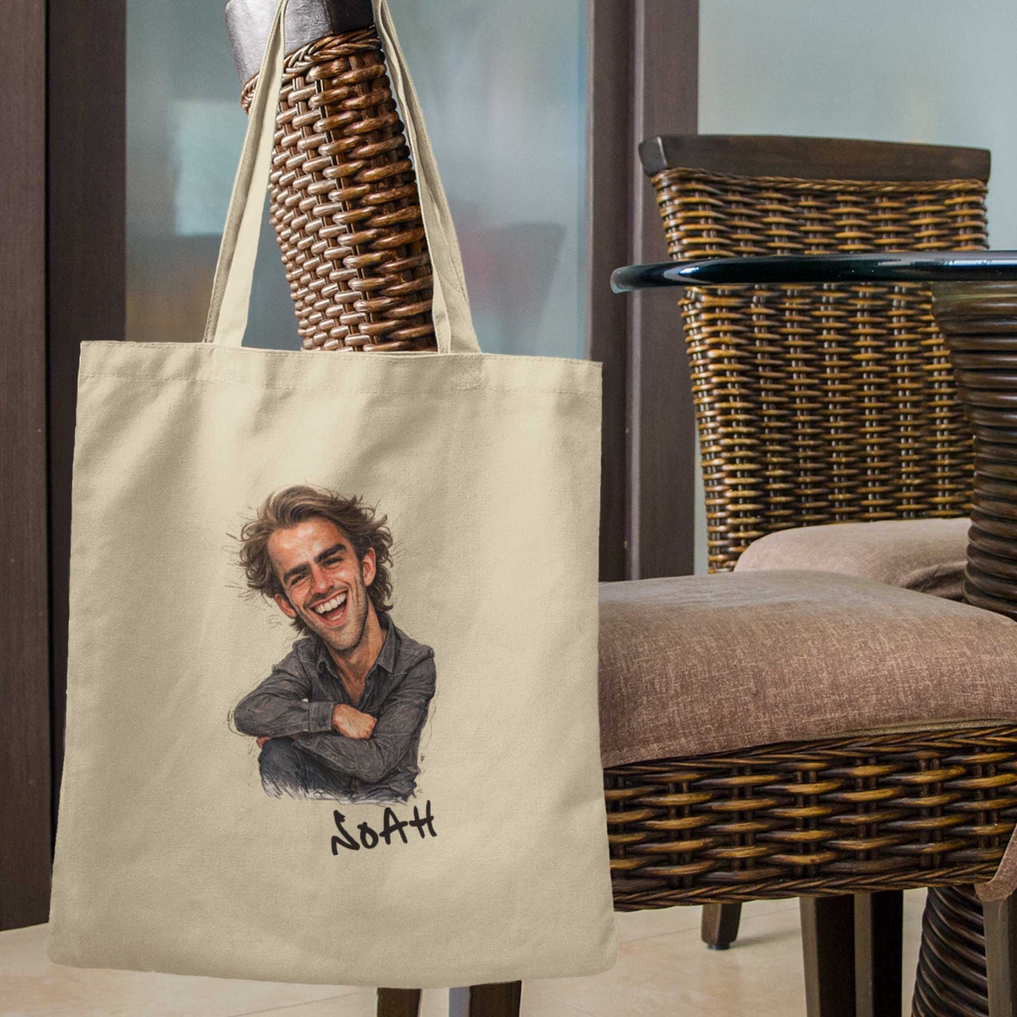 Beige katoenen totebag met gepersonaliseerde karikatuur van een lachende man met warrig haar en de naam Noah onder de illustratie, hangend aan een rotan stoel op een sfeervolle buitenlocatie met gevlochten meubels op de achtergrond.