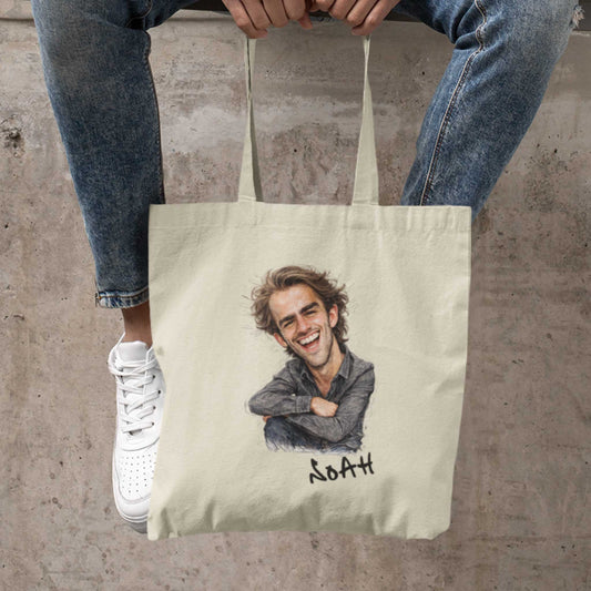Beige katoenen totebag met gepersonaliseerde karikatuur van een lachende man met warrig haar en de naam Noah onder de illustratie, vastgehouden tussen iemands benen terwijl die op een betonnen rand zit met jeans en witte sneakers zichtbaar in beeld.