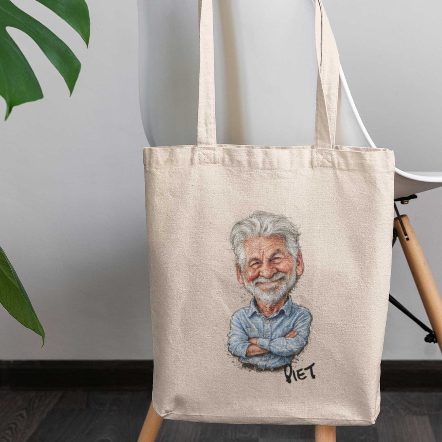 Beige katoenen totebag met gepersonaliseerde karikatuur van een oudere man met grijs haar en baard, inclusief de naam Piet onder de illustratie, geplaatst op een houten stoel in een modern interieur met lichte muur en groene bladeren op de achtergrond.