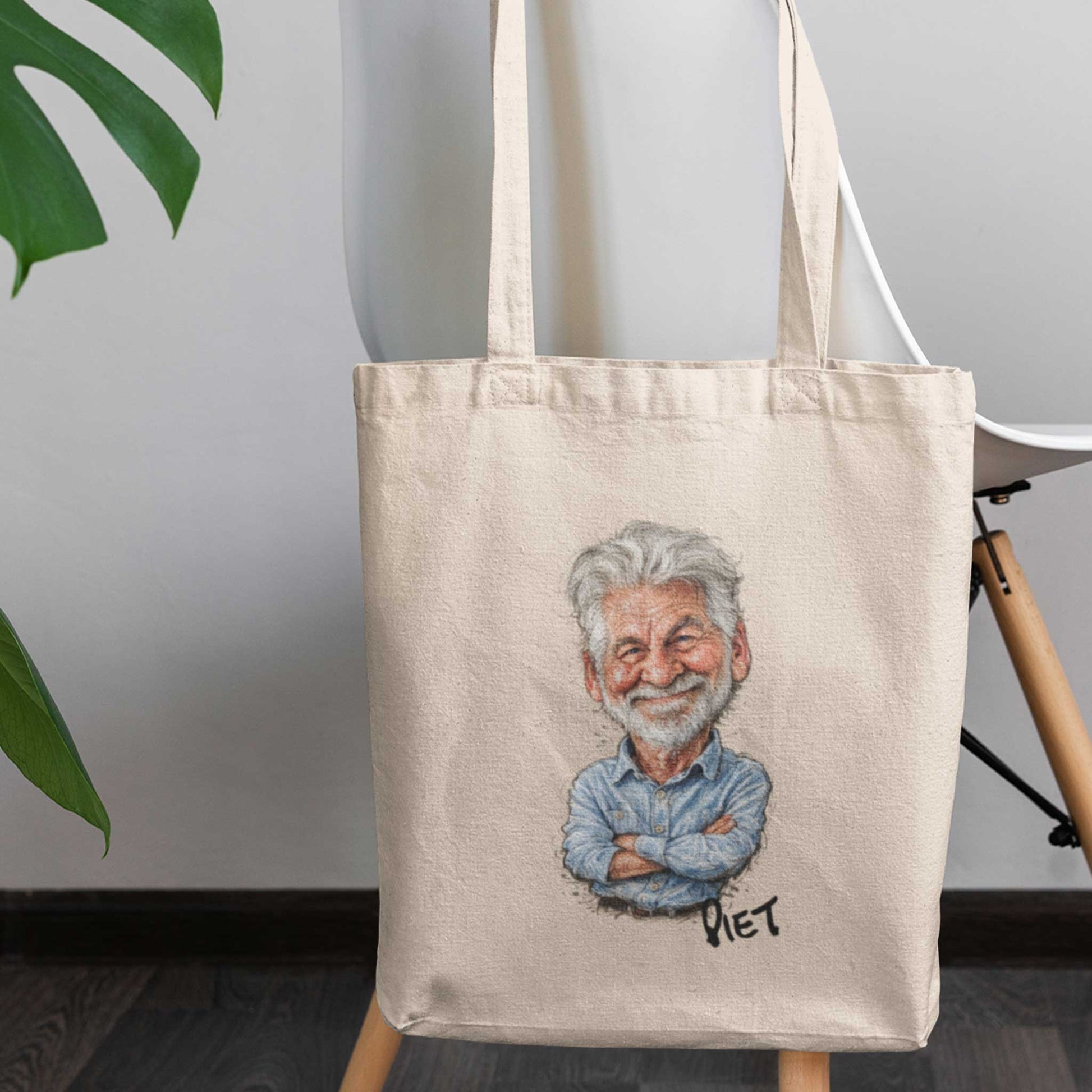 Beige katoenen totebag met gepersonaliseerde karikatuur van een oudere man met grijs haar en baard, inclusief de naam Piet onder de illustratie, geplaatst op een houten stoel in een modern interieur met lichte muur en groene bladeren op de achtergrond.