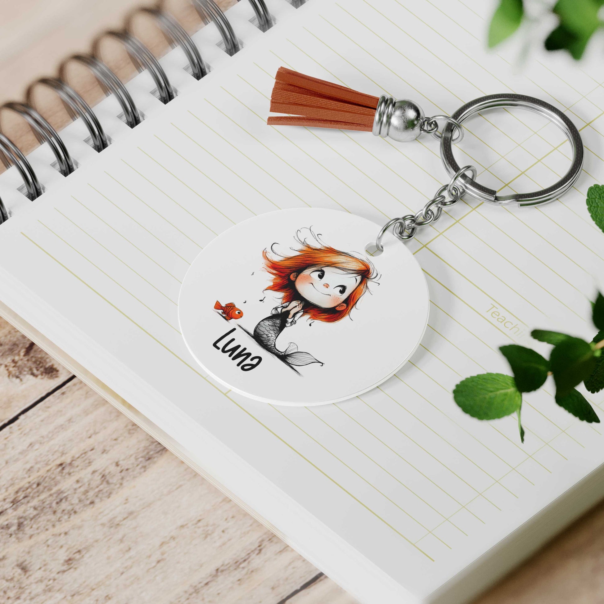 Gepersonaliseerde zeemeermin sleutelhanger met visje en voorbeeldnaam Luna liggend op een open notitieboek naast groene bladeren.