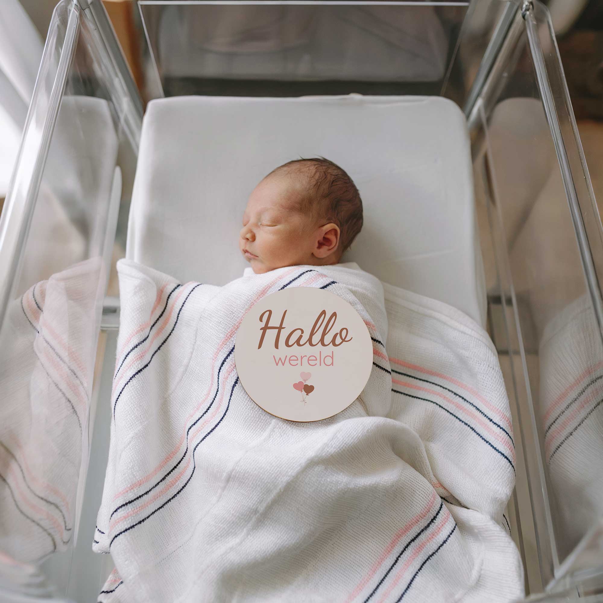 Pasgeboren baby liggend in een ziekenhuiswiegje met een ‘Hallo wereld’ mijlpaalschijfje op de deken als eerste fotomoment na de geboorte