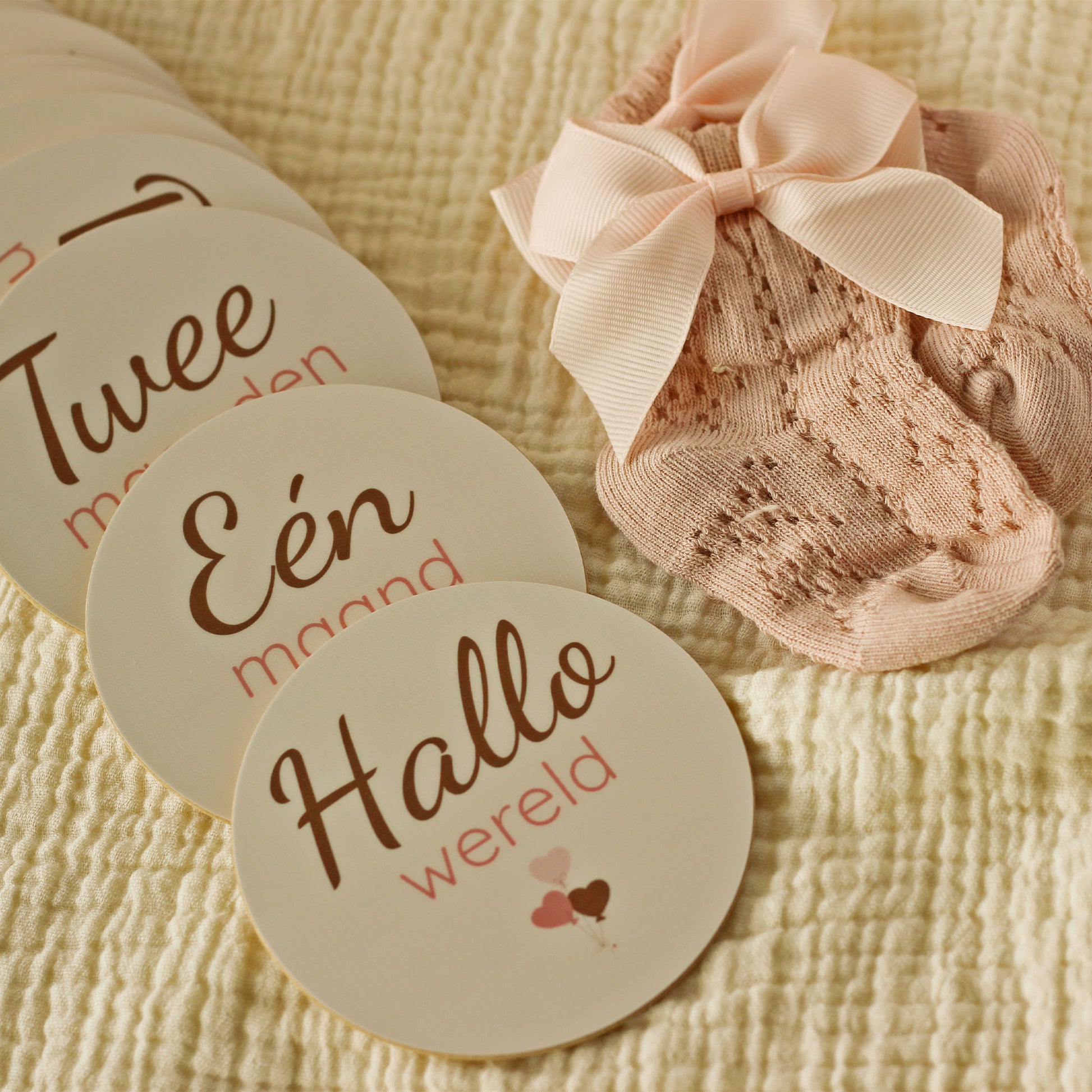 Close-up van baby mijlpaalschijfjes met teksten ‘Hallo wereld’, ‘Één maand’ en ‘Twee maanden’ naast roze babysokjes met strik, liggend op een zachte beige hydrofiele ondergrond in warme neutrale tinten