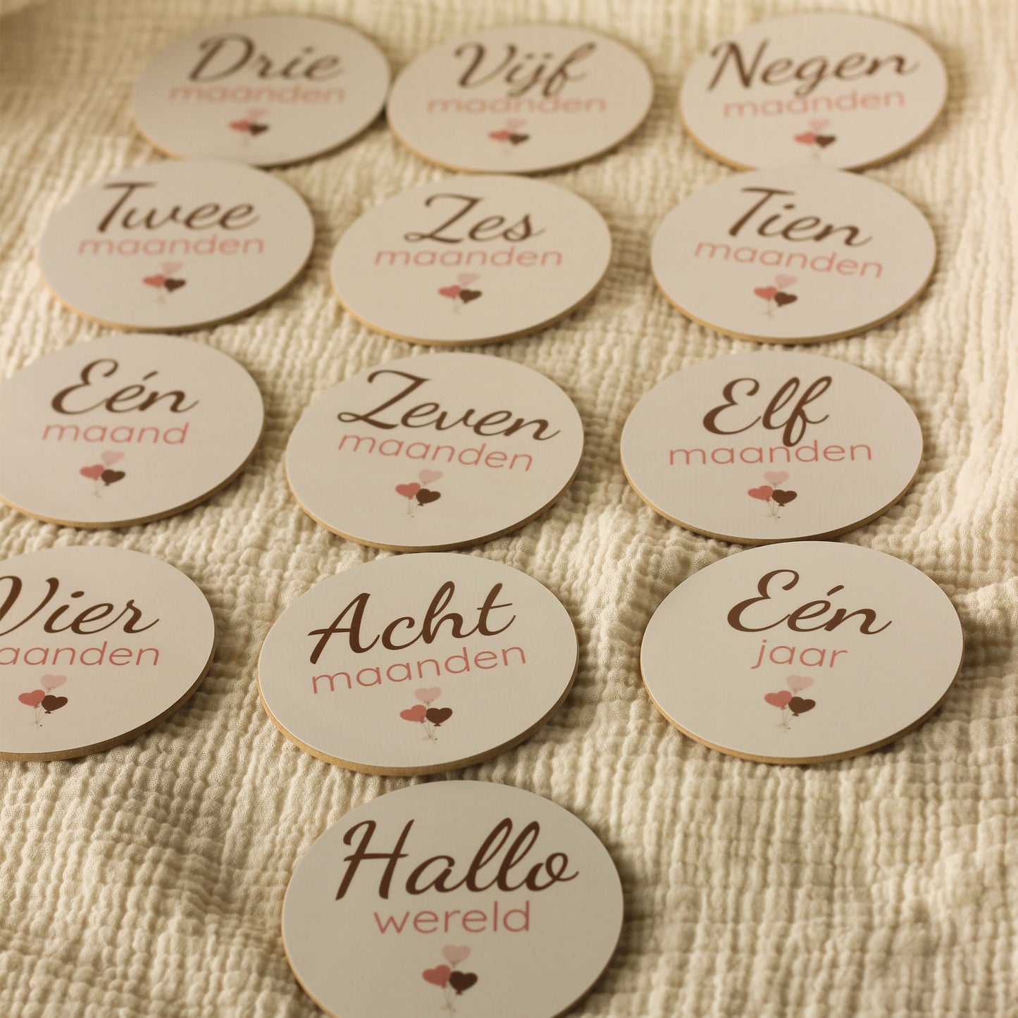 Complete set baby mijlpaalschijfjes voor het eerste levensjaar met ronde houten schijfjes voorzien van maand aanduidingen van één maand tot één jaar en ‘Hallo wereld’, netjes uitgestald op een zachte beige hydrofiele ondergrond