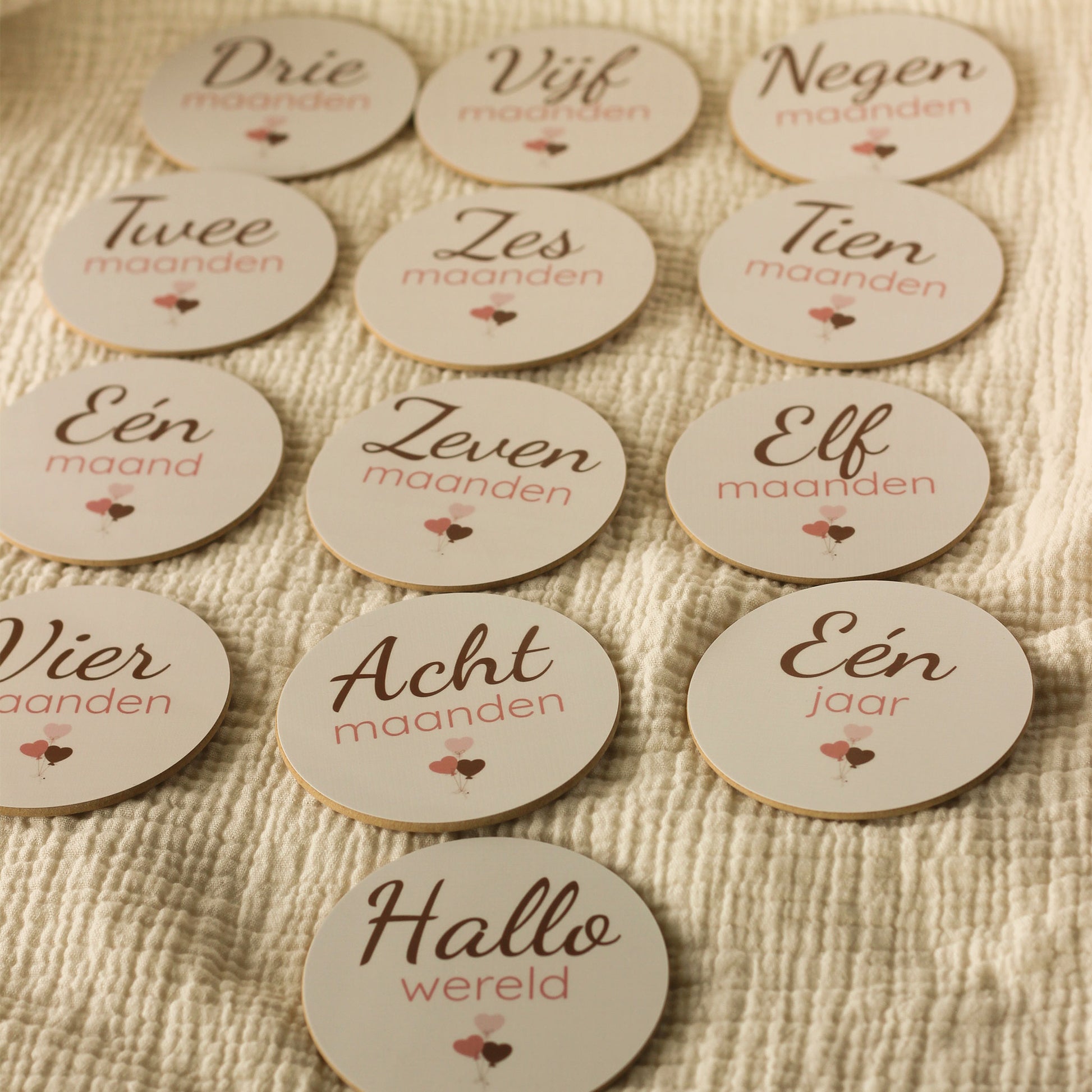 Complete set baby mijlpaalschijfjes voor het eerste levensjaar met ronde houten schijfjes voorzien van maand aanduidingen van één maand tot één jaar en ‘Hallo wereld’, netjes uitgestald op een zachte beige hydrofiele ondergrond