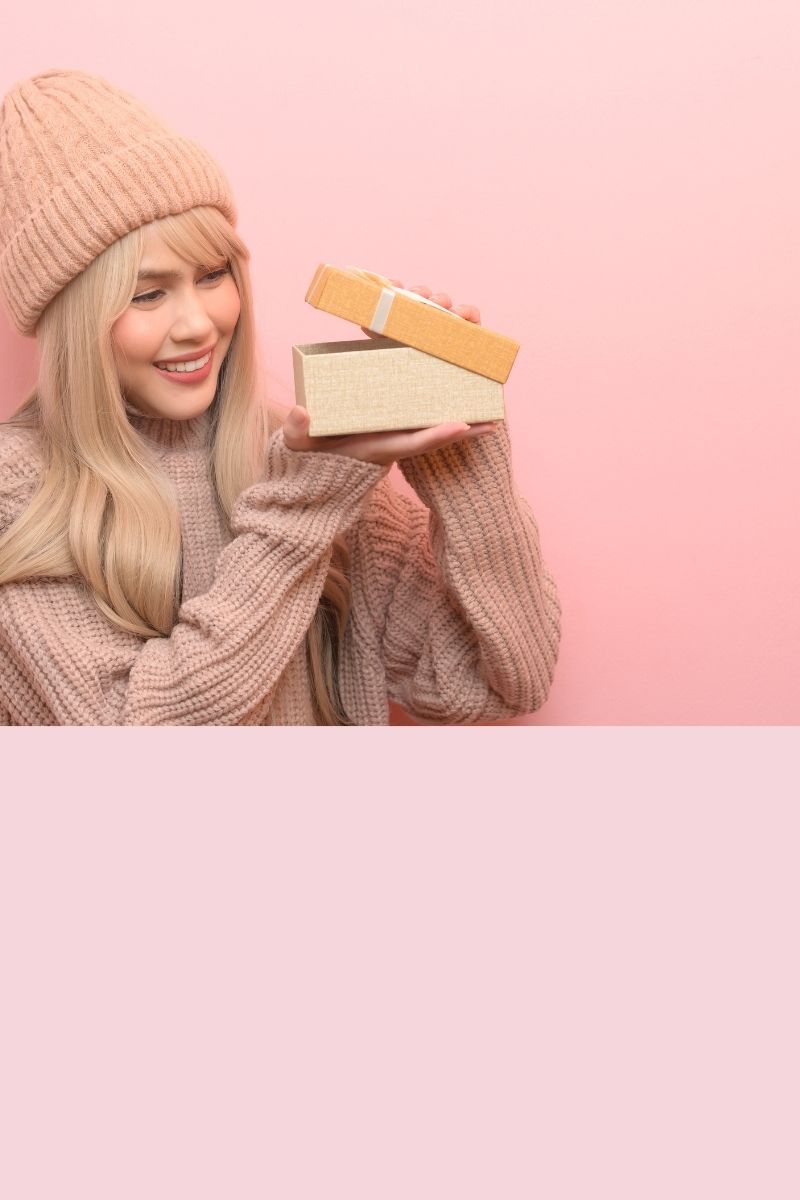 Blonde vrouw in roze trui opent een cadeau op een zachte roze achtergrond – perfect als header voor cadeaushop