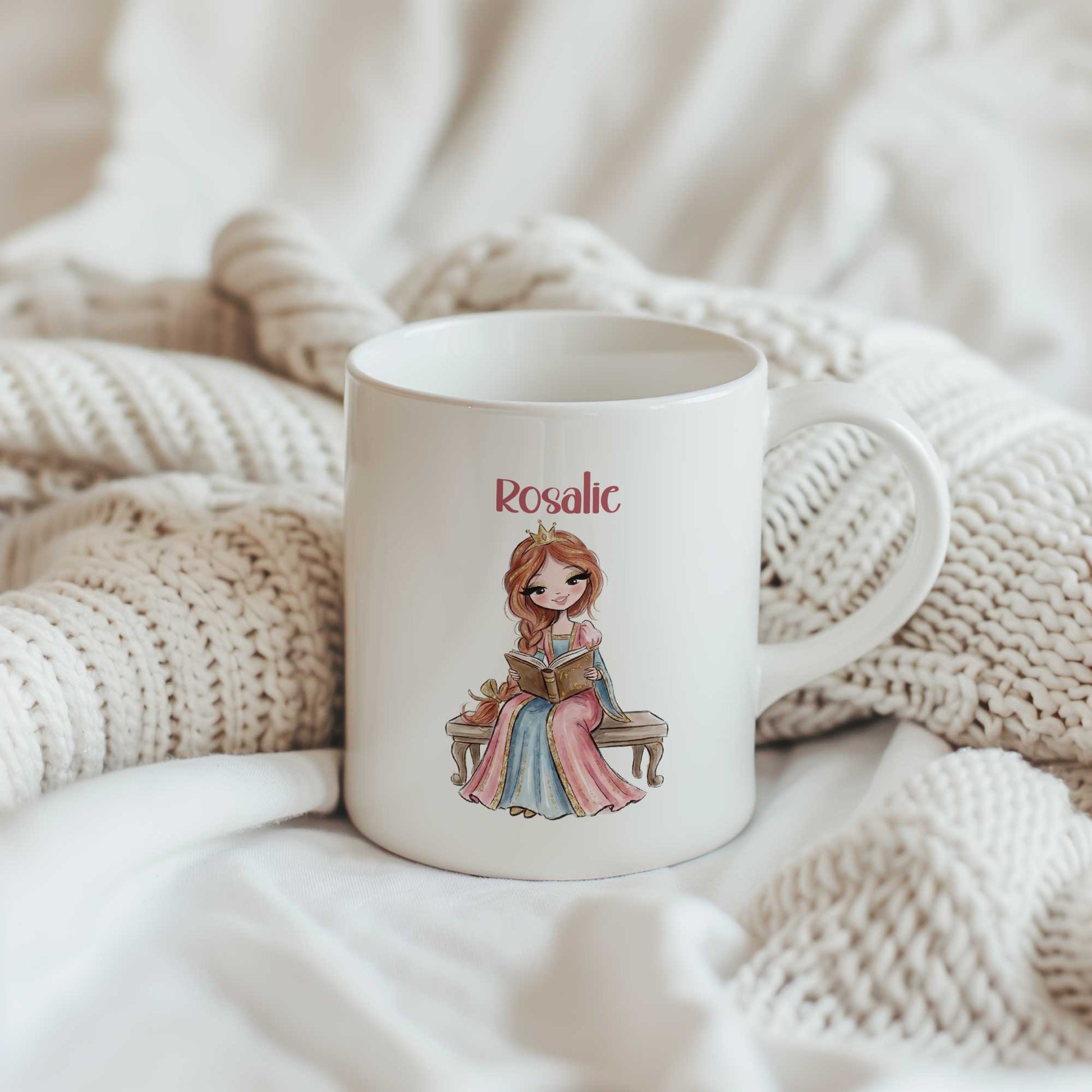 Witte mok tussen zachte gebreide dekens met een illustratie van een prinses op een bankje die een boek leest. Boven de illustratie staat de naam Rosalie.