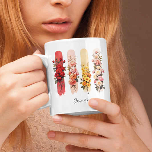 Vrouw met rood haar houdt een mok vast met rode rozen en kamperfoelie op gekleurde brushstrepen, tekst ‘Juni’ zichtbaar.