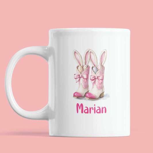 Witte mok met illustratie van roze westernlaarsjes gevuld met paaseieren en konijnenoren, met de naam Marian in speels roze lettertype, op een effen roze achtergrond.
