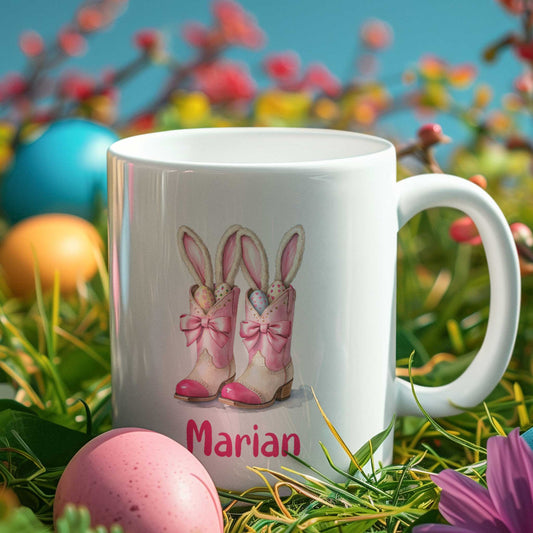 Witte mok met roze laarsjes gevuld met gekleurde paaseieren en konijnenoren, afgewerkt met strikken en de naam Marian, geplaatst tussen gras en paaseieren in vrolijke lentekleuren.

