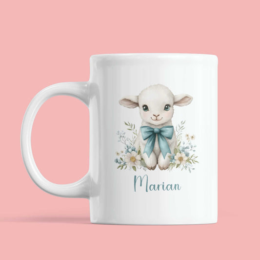 Witte mok met een aquarelillustratie van een wit lammetje met grote blauwe strik en bloemenkrans onderaan, met de naam Marian in sierlijk blauw schrift op een effen roze achtergrond.