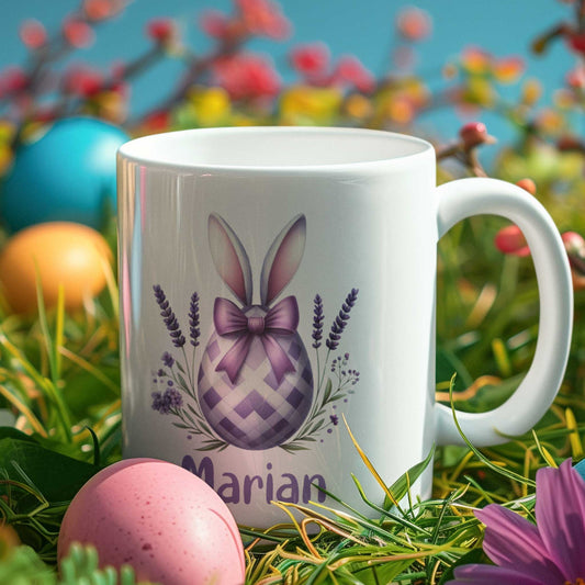 Witte mok met een paars ruitjes-paasei versierd met konijnenoren, lavendelbloemen en een grote paarse strik. Gepersonaliseerd met de naam Marian. De mok staat tussen gras, paaseieren en bloemen in een lenteachtige setting.