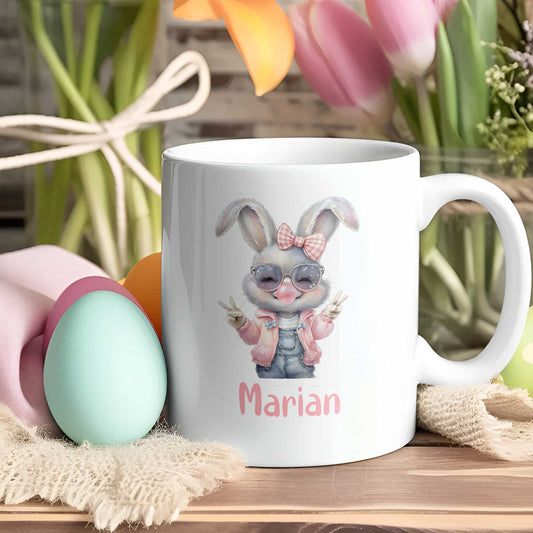 Witte mok met cool meisjeskonijn, zonnebril, kauwgombel en naam Marian in roze, tussen paaseieren en bloemen op houten tafel.
