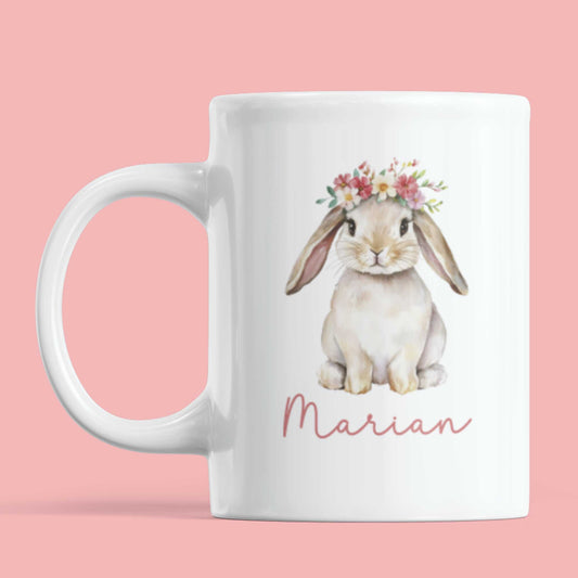 Witte keramieken mok met een schattig konijn met bloemenkrans en de naam Marian in sierlijk roze schrift, op een roze achtergrond.
