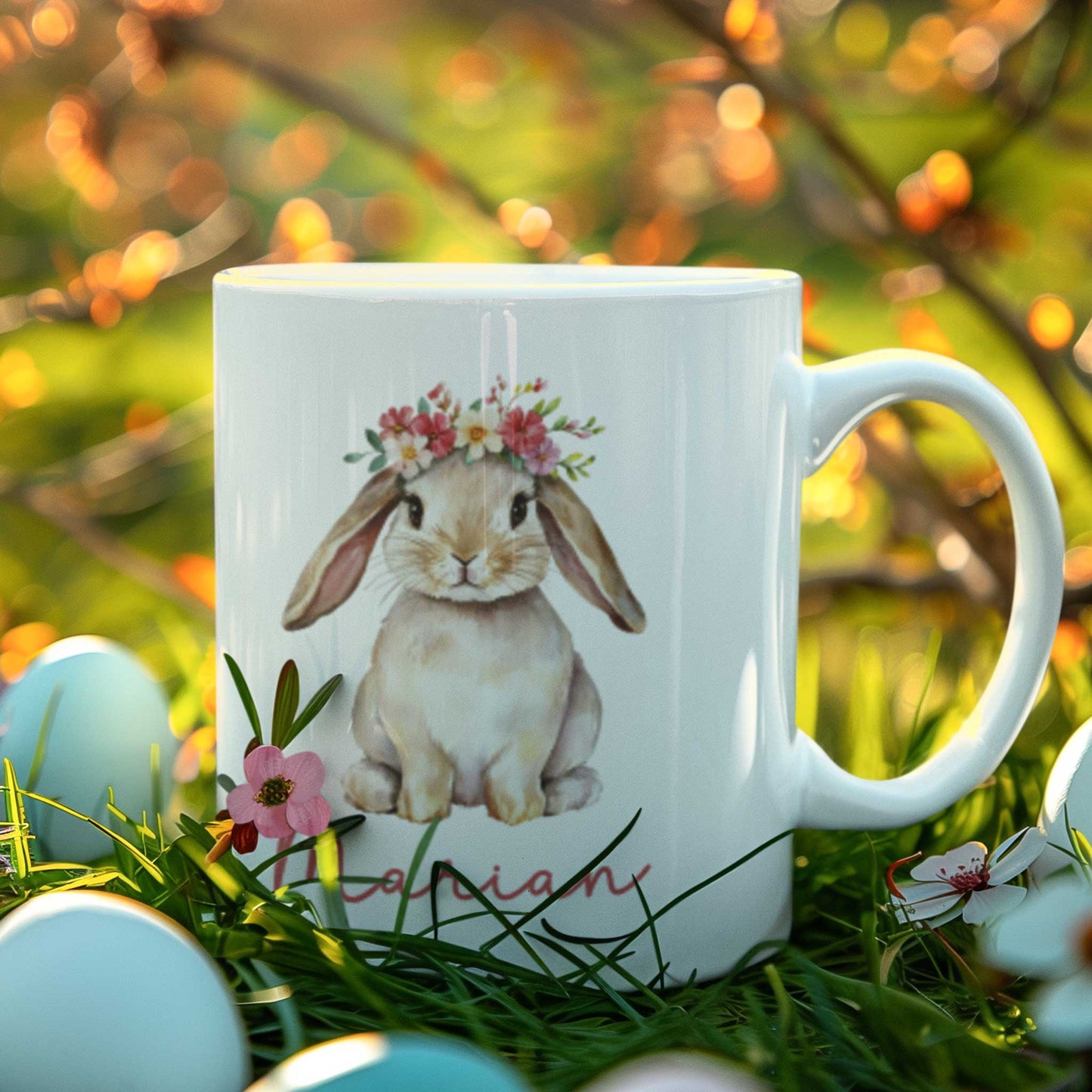 Witte mok met een illustratie van een bruin konijntje met bloemenkrans en de naam Marian, geplaatst in gras tussen gekleurde paaseieren.