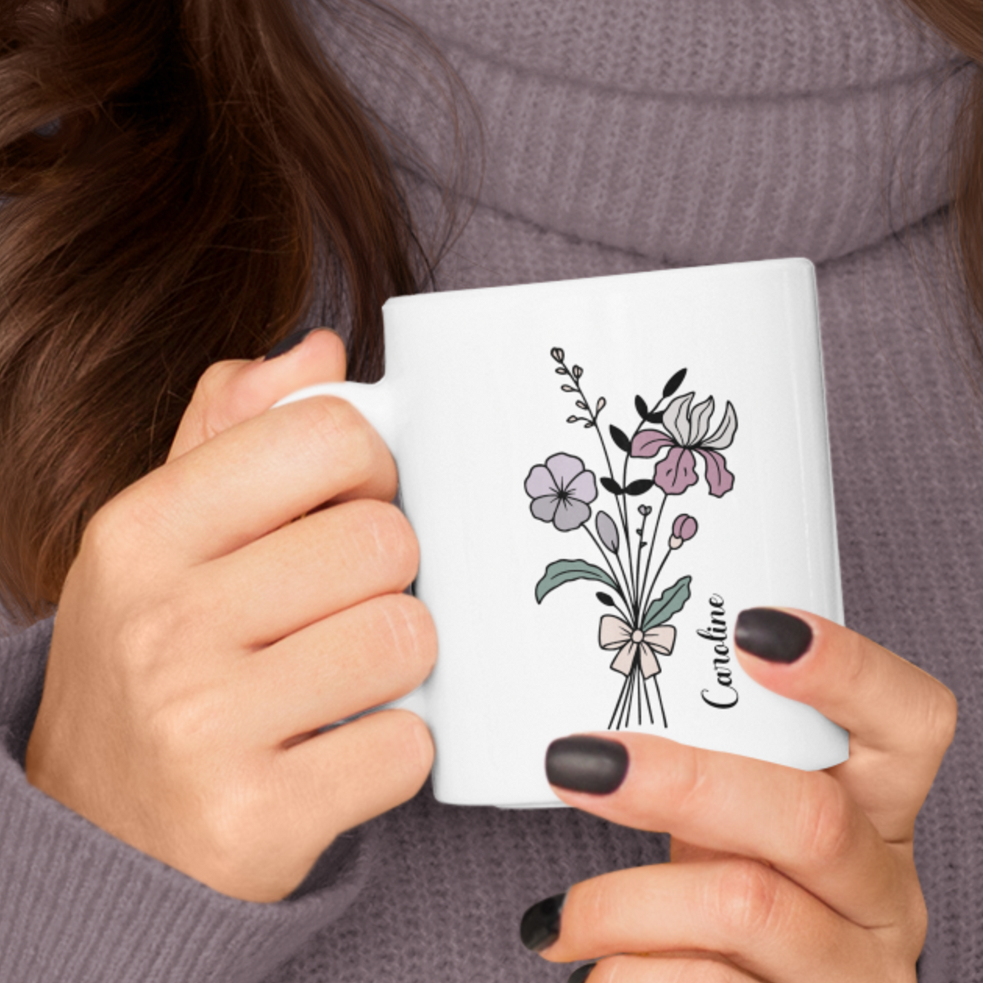 vrouw met paars grijze coltrui en een gepersonaliseerde witte mok met lijnbloemen in paars roze tinten en een sierlijke naam in het zwart in haar hand
