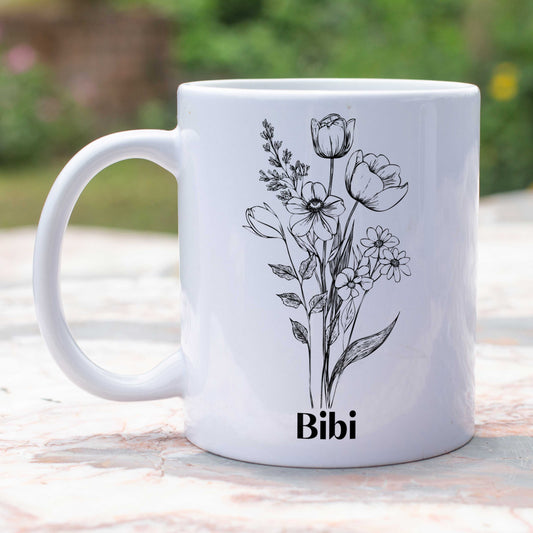 gepersonaliseerde witte mok met een zwarte schets van bloemen en een naam in zwart buiten in de tuin op een tuintafel