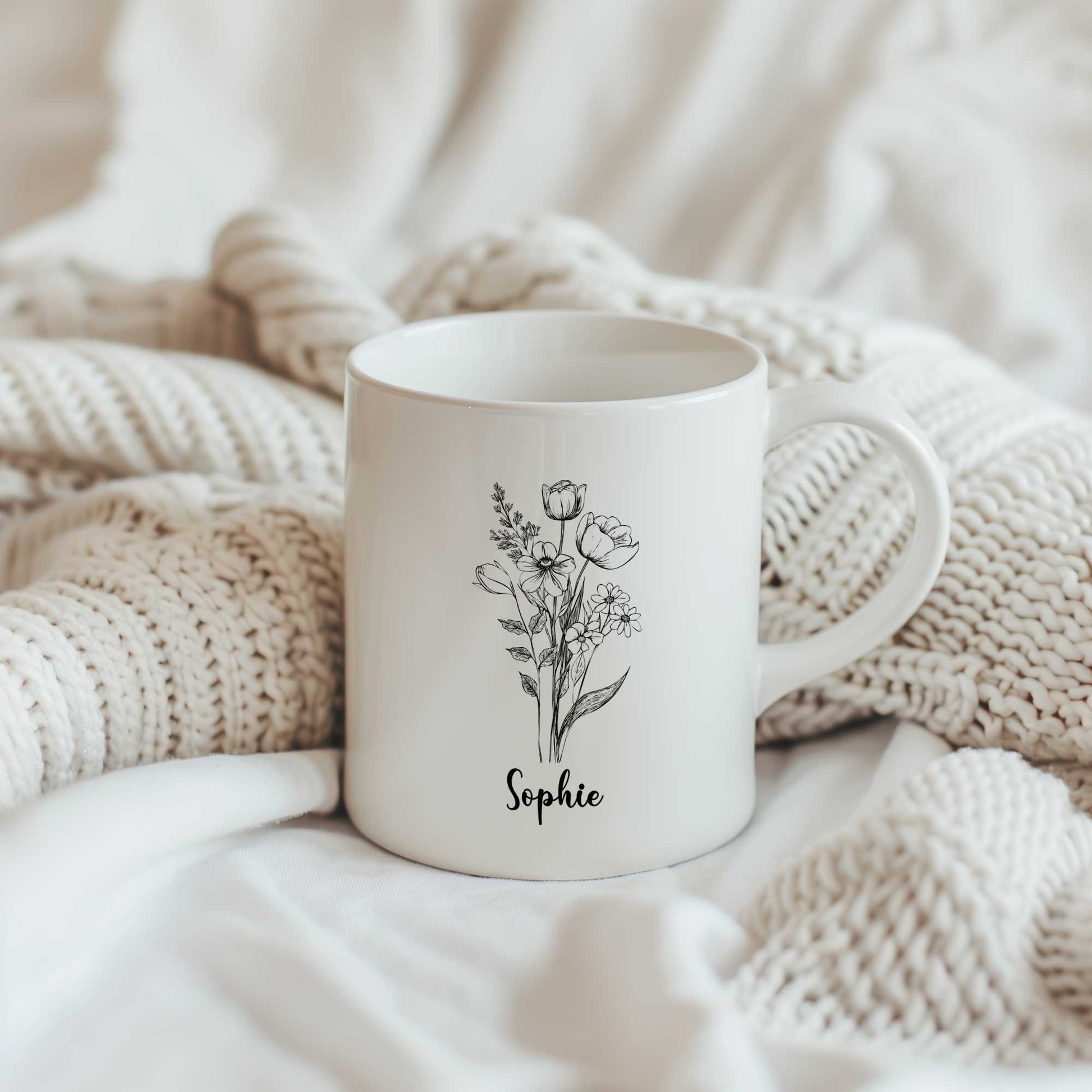 Sfeerfoto van een witte mok met een zwarte bloemenschets en de naam Sophie op een gebreide beige deken.