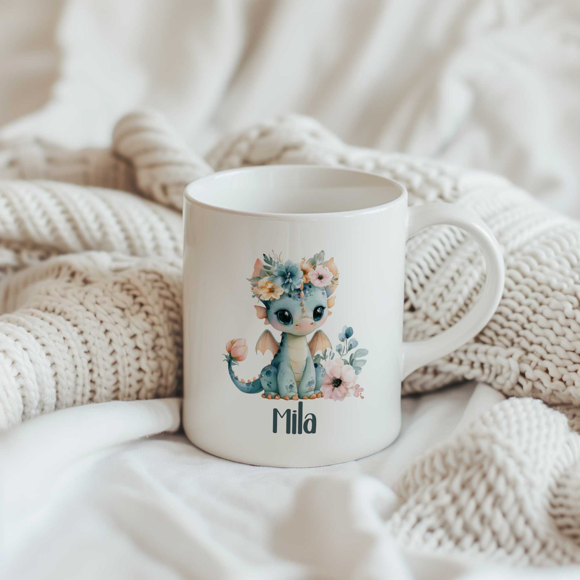 Witte mok met een schattig blauw draakje met bloemenkroon en de naam Mila, geplaatst tussen zachte gebreide dekens.