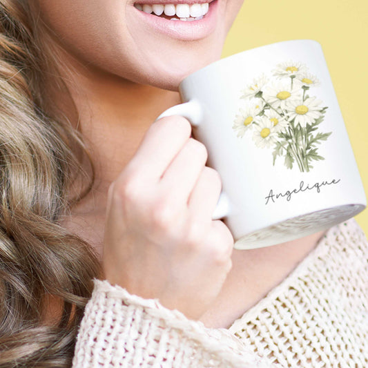 Vrouw drinkt uit witte mok met madeliefjes en de naam Angelique, tegen een gele achtergrond.