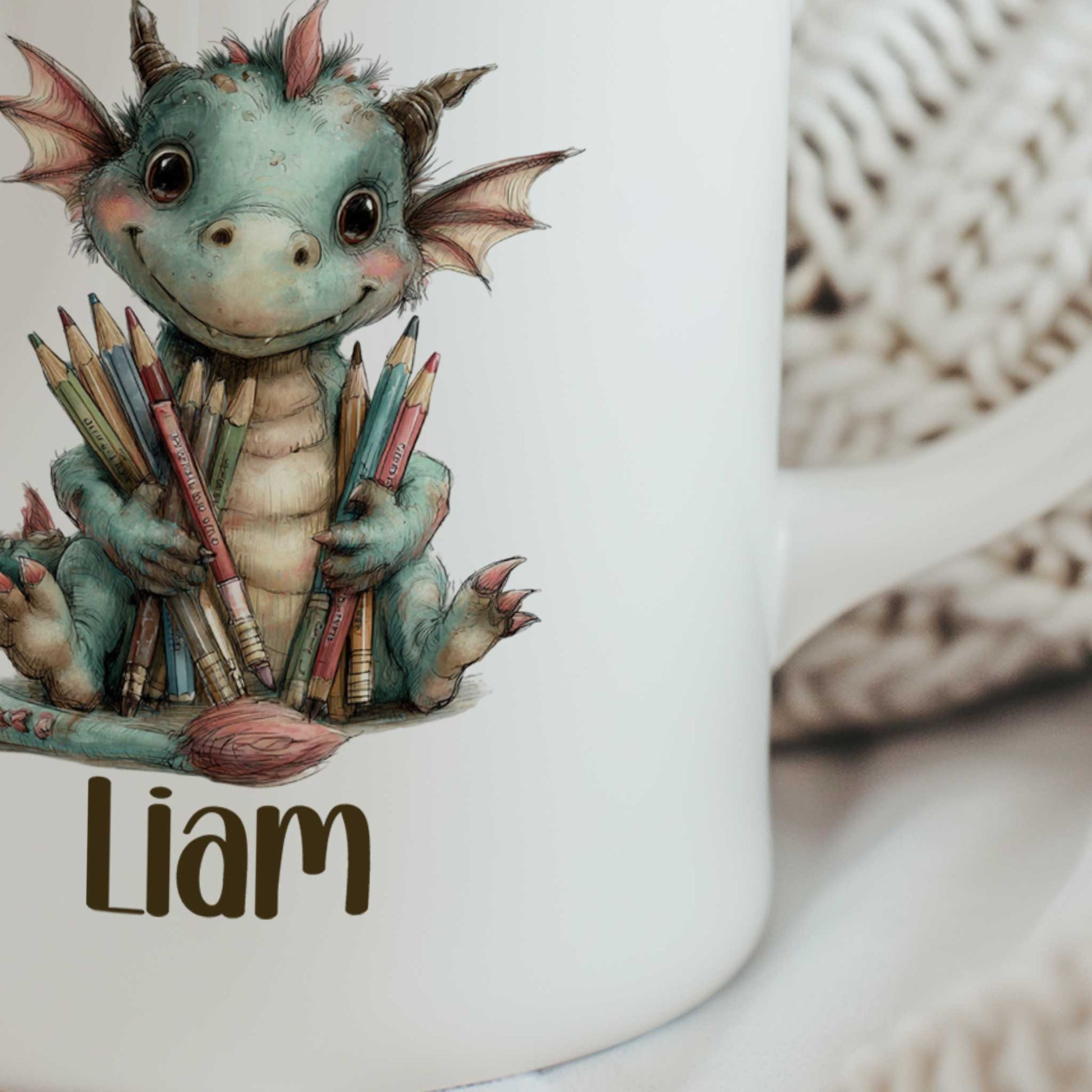 Close-up van een witte gepersonaliseerde mok met een vrolijk groen draakje dat kleurpotloden en penselen vasthoudt, met de naam Liam onder de illustratie.