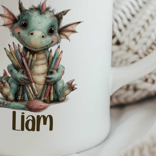 Close-up van een witte gepersonaliseerde mok met een vrolijk groen draakje dat kleurpotloden en penselen vasthoudt, met de naam Liam onder de illustratie.