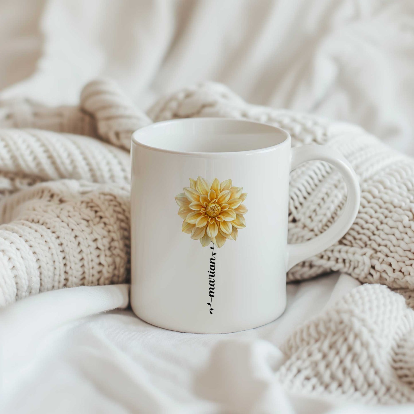 Witte keramische mok met een gele dahlia waarvan de bloemsteel wordt gevormd door een gepersonaliseerde naam, geplaatst tussen zachte crèmekleurige gebreide dekens.