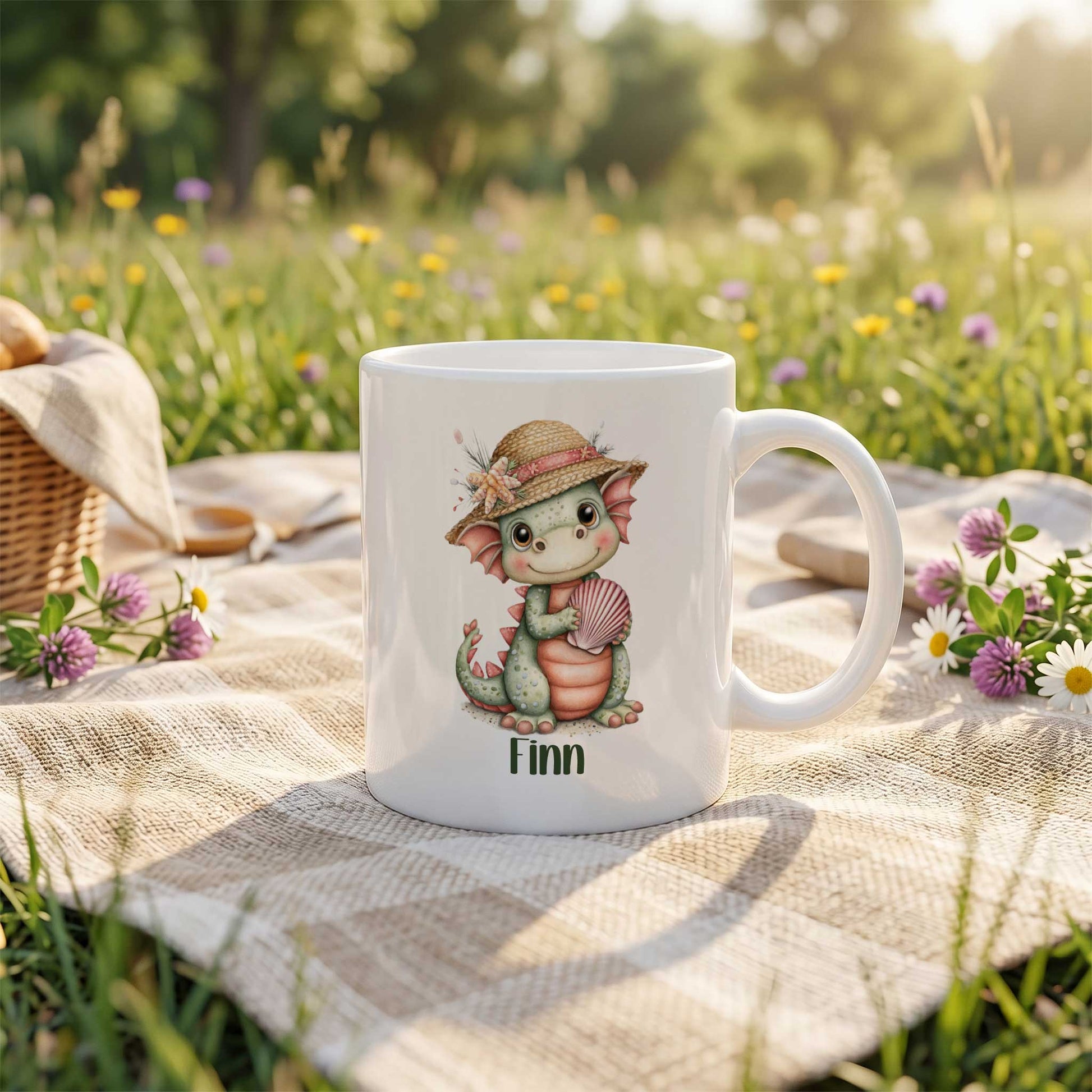 Witte mok met een groen draakje met een strohoedje en een roze schelp. Onder de illustratie staat de naam Finn. De mok staat buiten op een kleed tussen gras en bloemen.