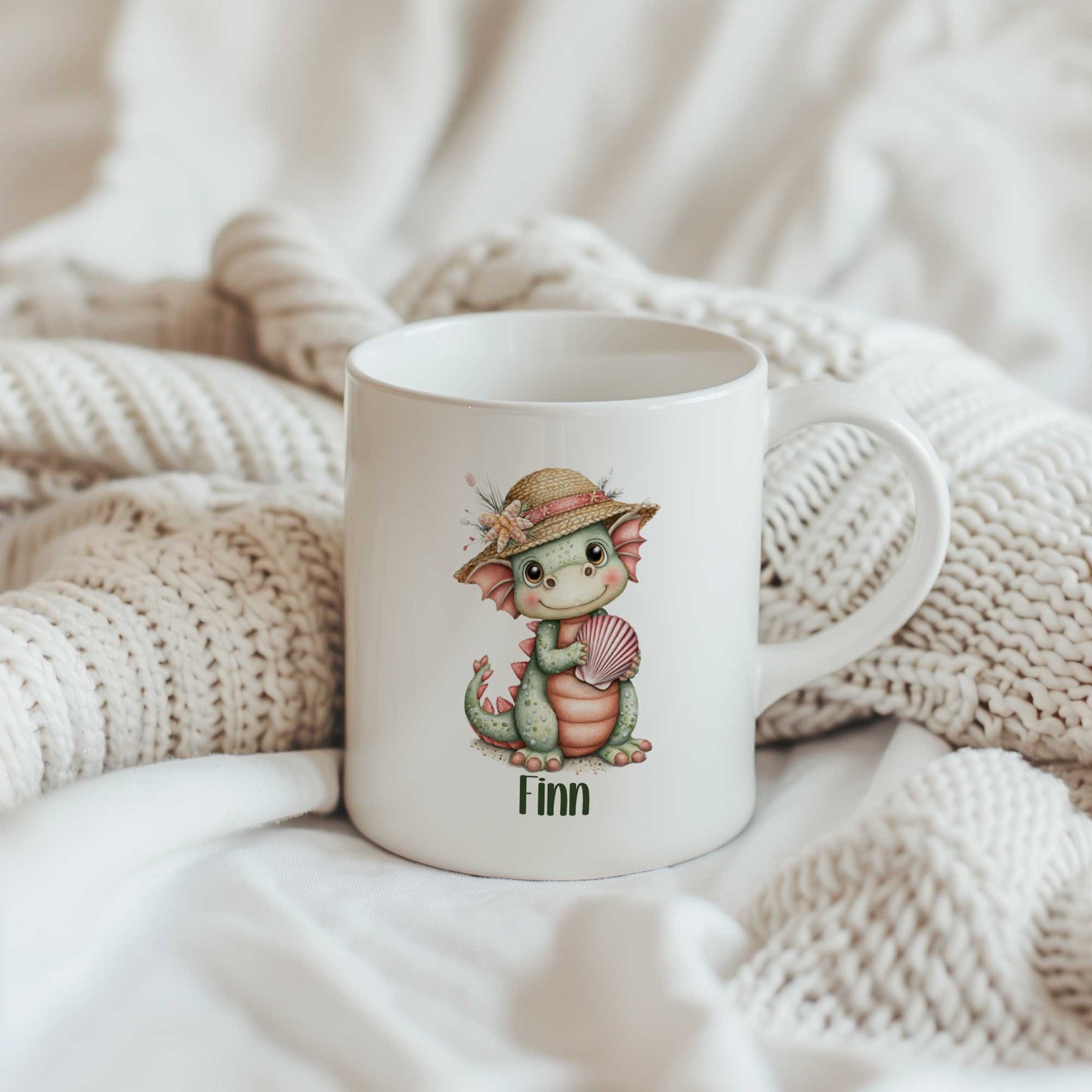 Witte mok met een groen draakje met strohoedje en roze schelp. Onder de illustratie staat de naam Finn. De mok staat tussen beige gebreide dekens.