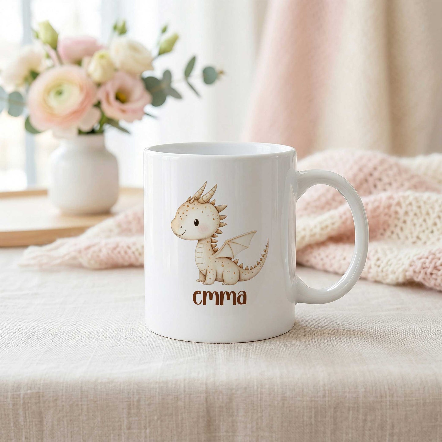 Witte mok met beige draakje Vonkje en de naam Emma op een lichte tafel naast een zachtroze plaid en een vaas met bloemen.