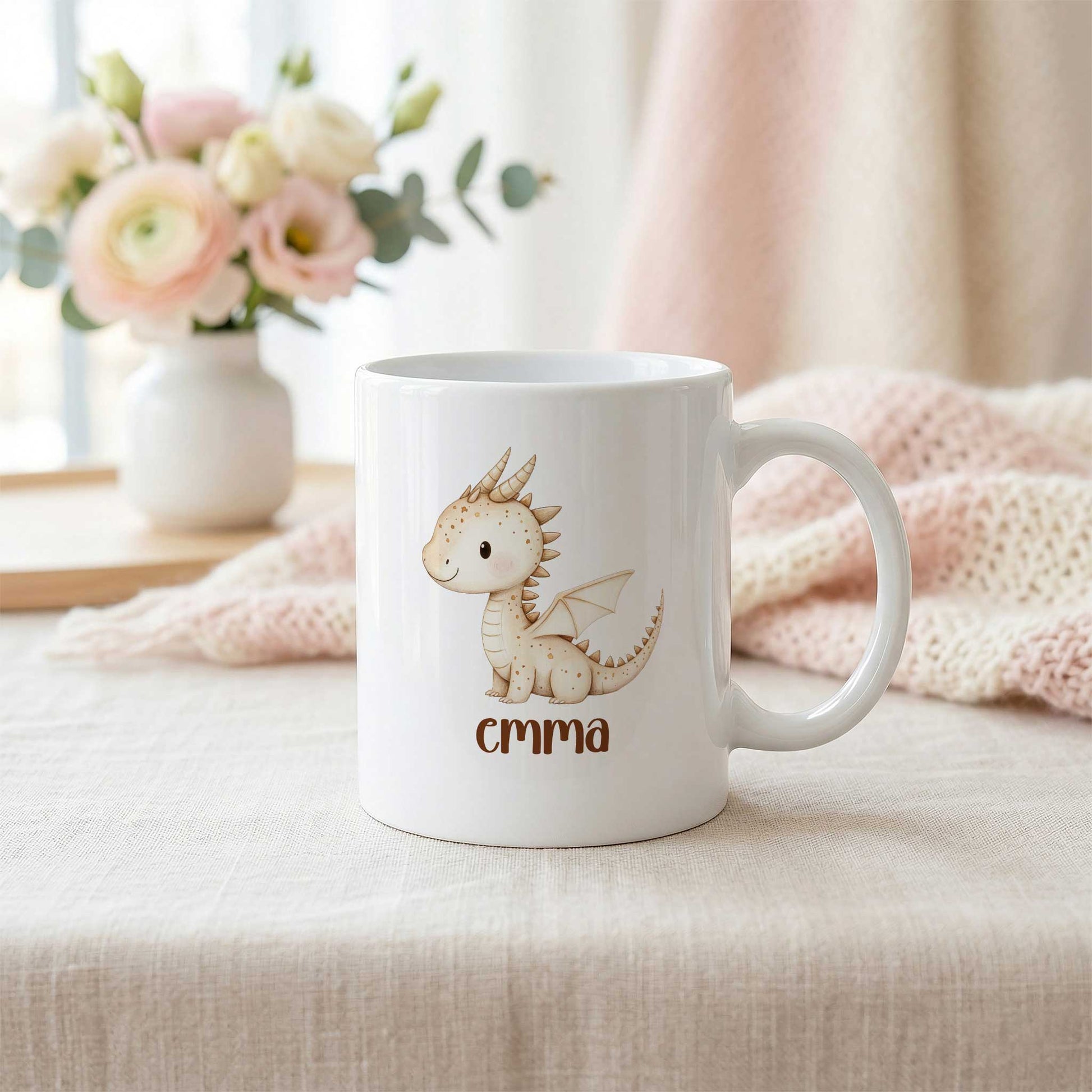 Witte mok met beige draakje Vonkje en de naam Emma op een lichte tafel naast een zachtroze plaid en een vaas met bloemen.