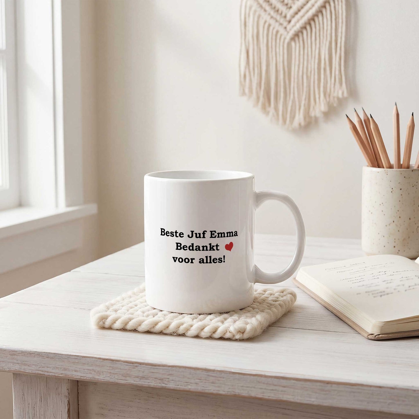 Witte keramische mok bedrukt met de tekst 'Beste Juf Emma Bedankt voor alles!' en een rood hartje, geplaatst op een licht houten bureau naast een notitieboek