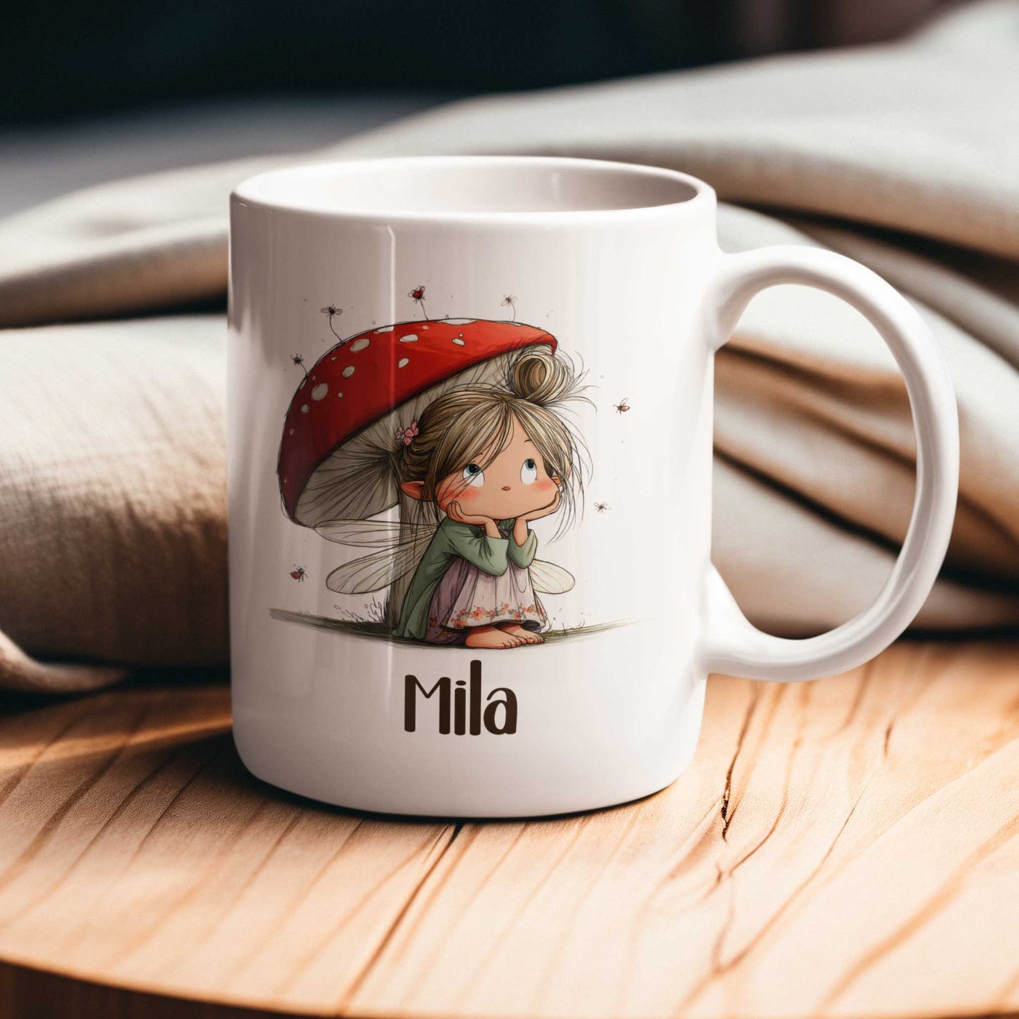 Witte mok met fee-illustratie en naam Mila op een houten tafel met beige stoffen op de achtergrond.