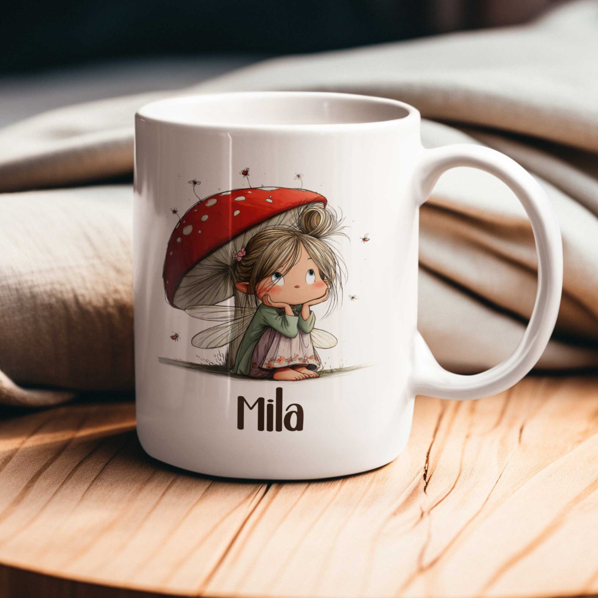 Witte mok met fee-illustratie en naam Mila op een houten tafel met beige stoffen op de achtergrond.