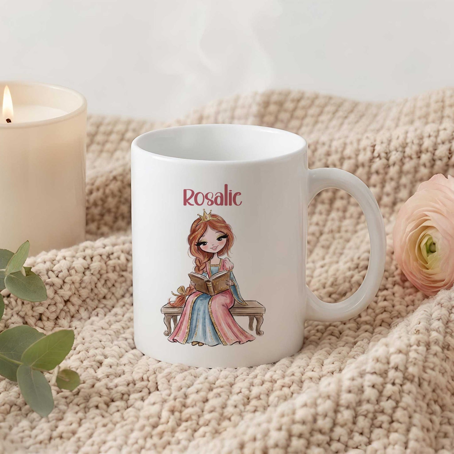 Witte mok op een beige gebreide deken naast een brandende kaars en een roze bloem. Op de mok staat een prinses op een bankje met een open boek en de naam Rosalie.