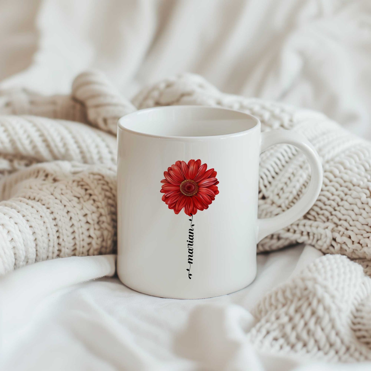 Witte keramische mok met een rode gerbera en gepersonaliseerde naam verwerkt in de bloemsteel, geplaatst op een beige gebreide deken.