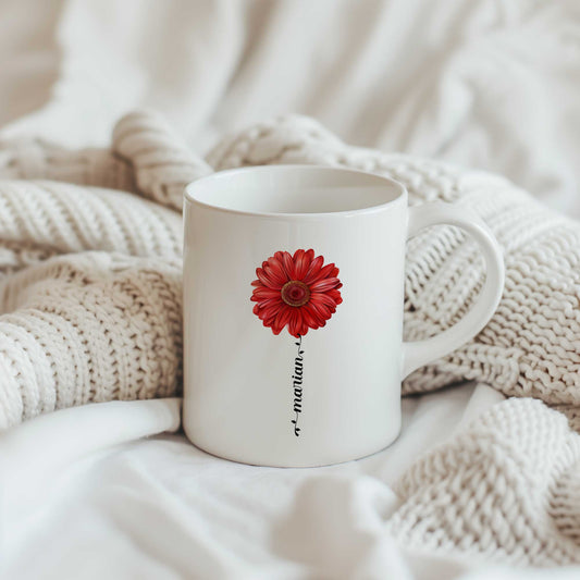Witte keramische mok met een rode gerbera en gepersonaliseerde naam verwerkt in de bloemsteel, geplaatst op een beige gebreide deken.