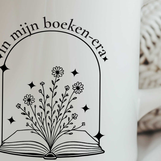 Close-up van een witte mok met een zwart ontwerp van een open boek waar bloemen uit groeien en de tekst ‘In mijn boeken-era’.