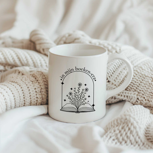 Witte mok met een open boek, bloemen en de tekst ‘In mijn boeken-era’, staand tussen beige gebreide dekens op een bed.