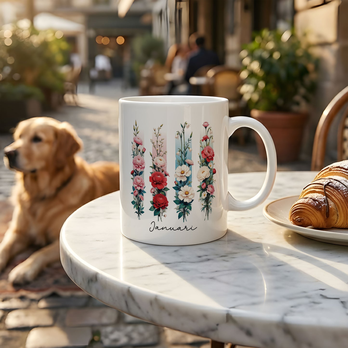 Witte mok met de geboortebloem van januari in brush effect op een marmeren terrastafel, met een golden retriever op de achtergrond en een bord met croissants ernaast.