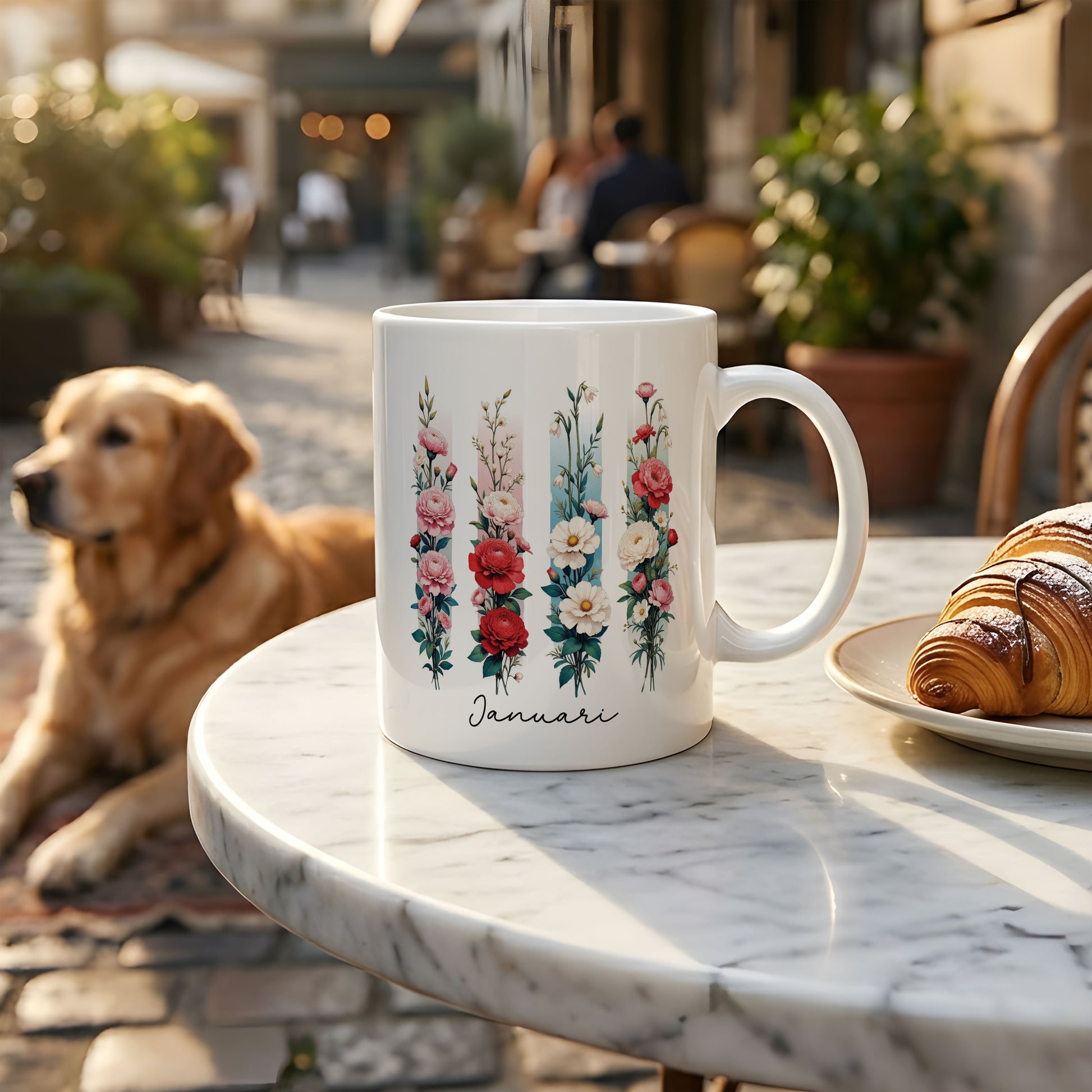Witte mok met de geboortebloem van januari in brush effect op een marmeren terrastafel, met een golden retriever op de achtergrond en een bord met croissants ernaast.