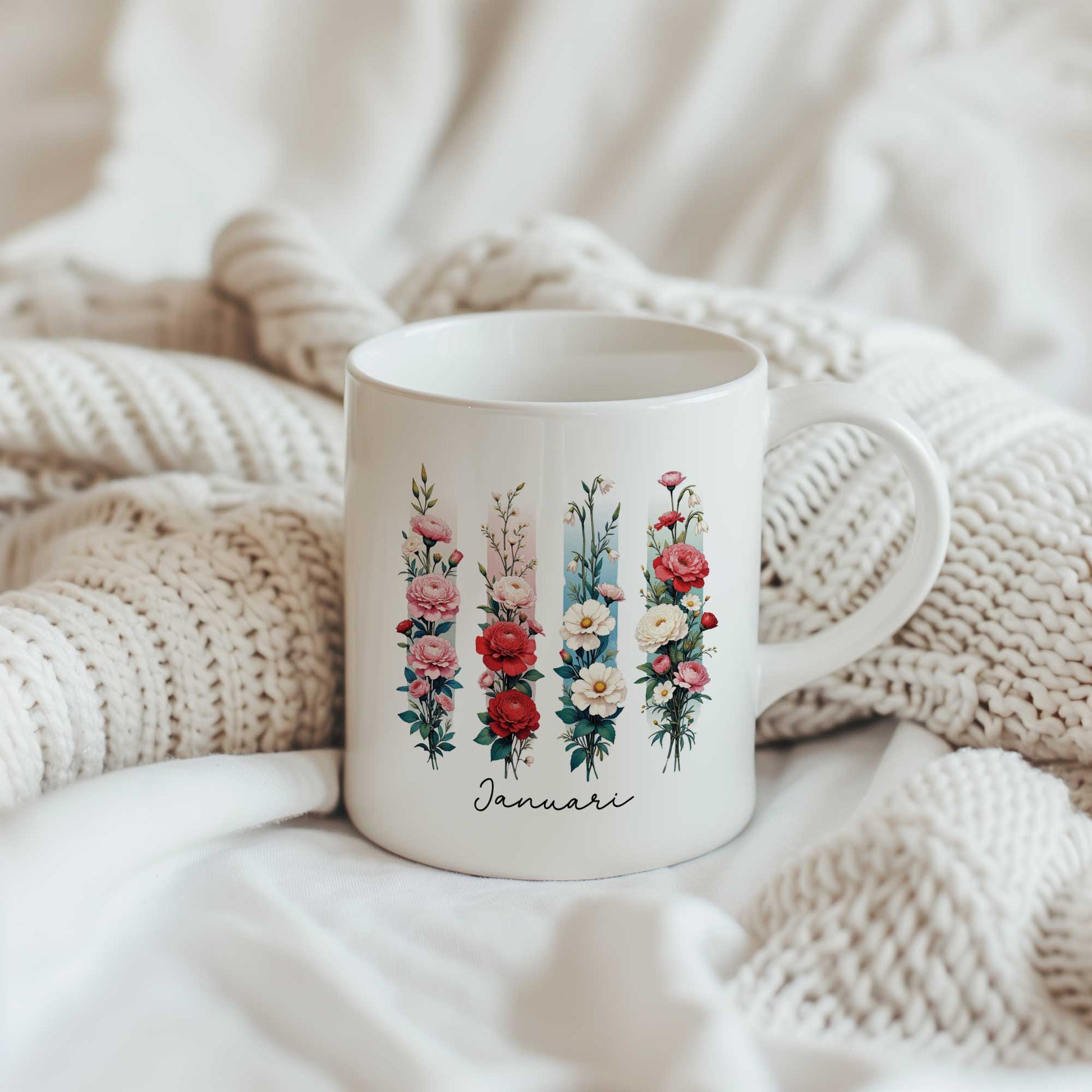 Witte mok met de geboortebloem van januari in brush effect op een beige gebreide plaid, met het bloemenontwerp en de tekst 'Januari' zichtbaar.