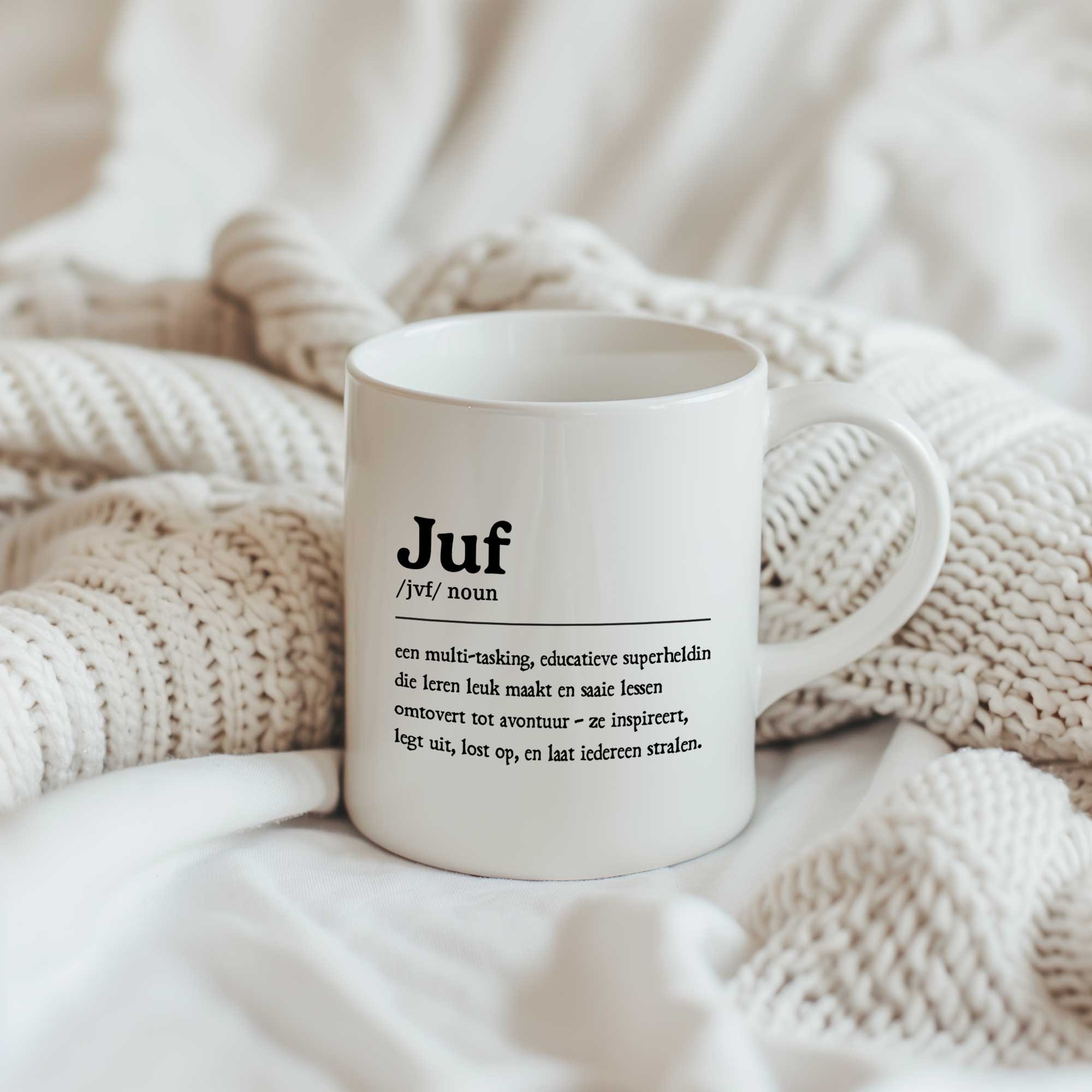 Witte keramische mok bedrukt met een definitie van een juf in woordenboekstijl. De mok staat op zachte crèmekleurige dekens.