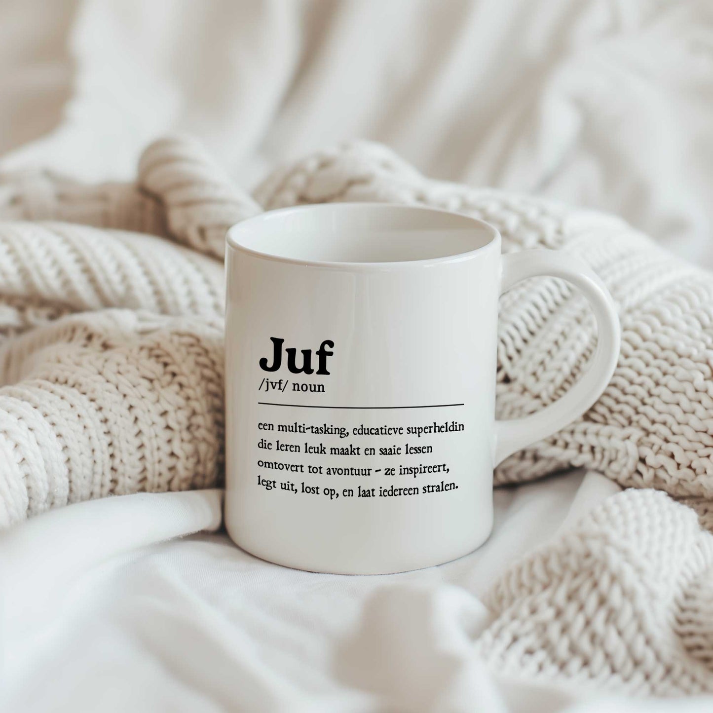 Witte keramische mok bedrukt met een definitie van een juf in woordenboekstijl. De mok staat op zachte crèmekleurige dekens.