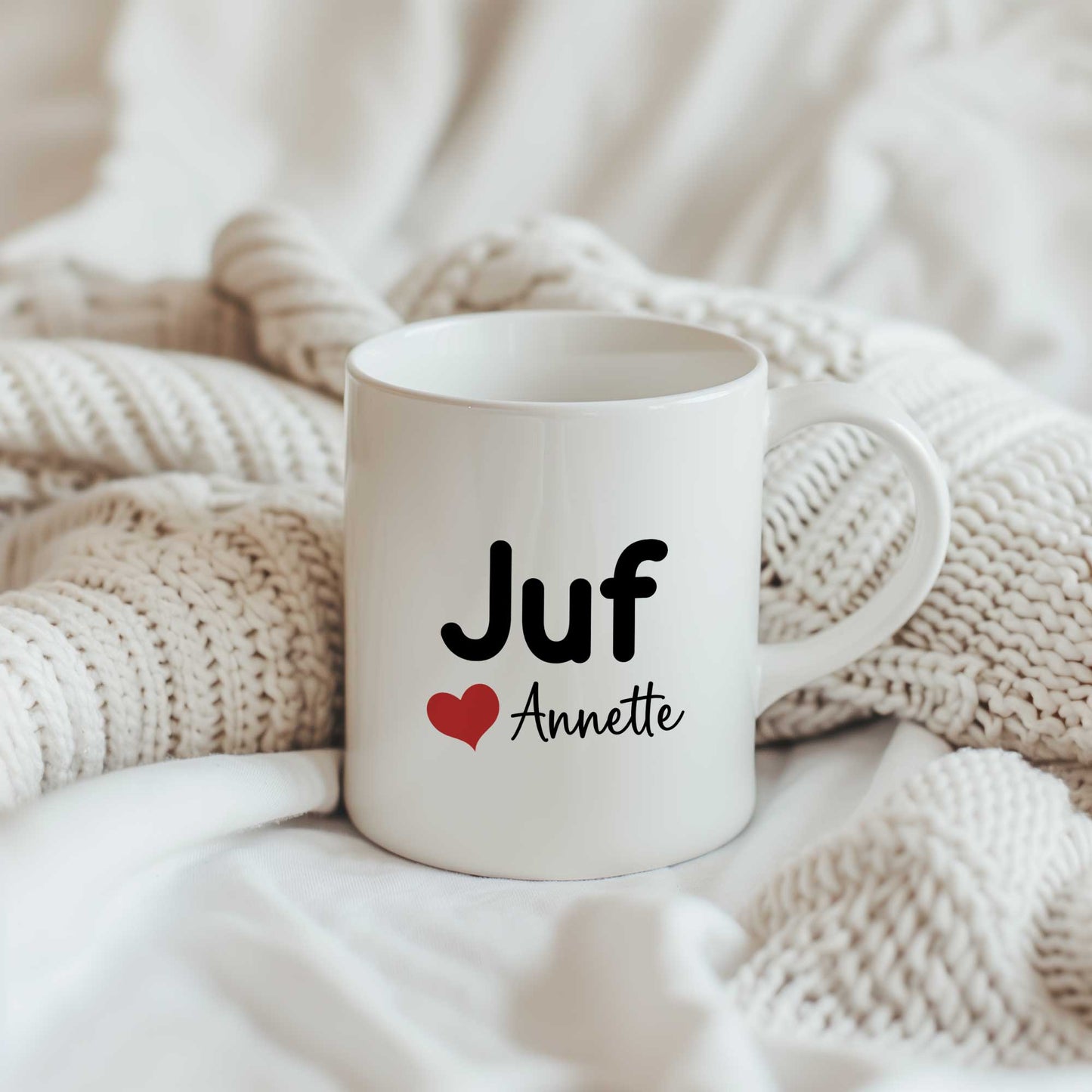 Sfeerfoto van een witte mok met de tekst Juf, een rood hartje en de naam Annette op een lichte gebreide deken