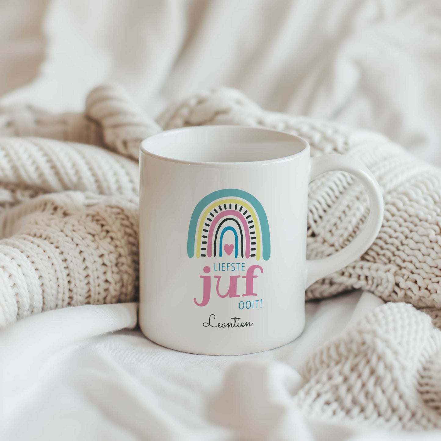 Witte mok met pastel regenboog, tekst 'Liefste juf ooit!' en gepersonaliseerde naam, geplaatst tussen beige gebreide dekens
