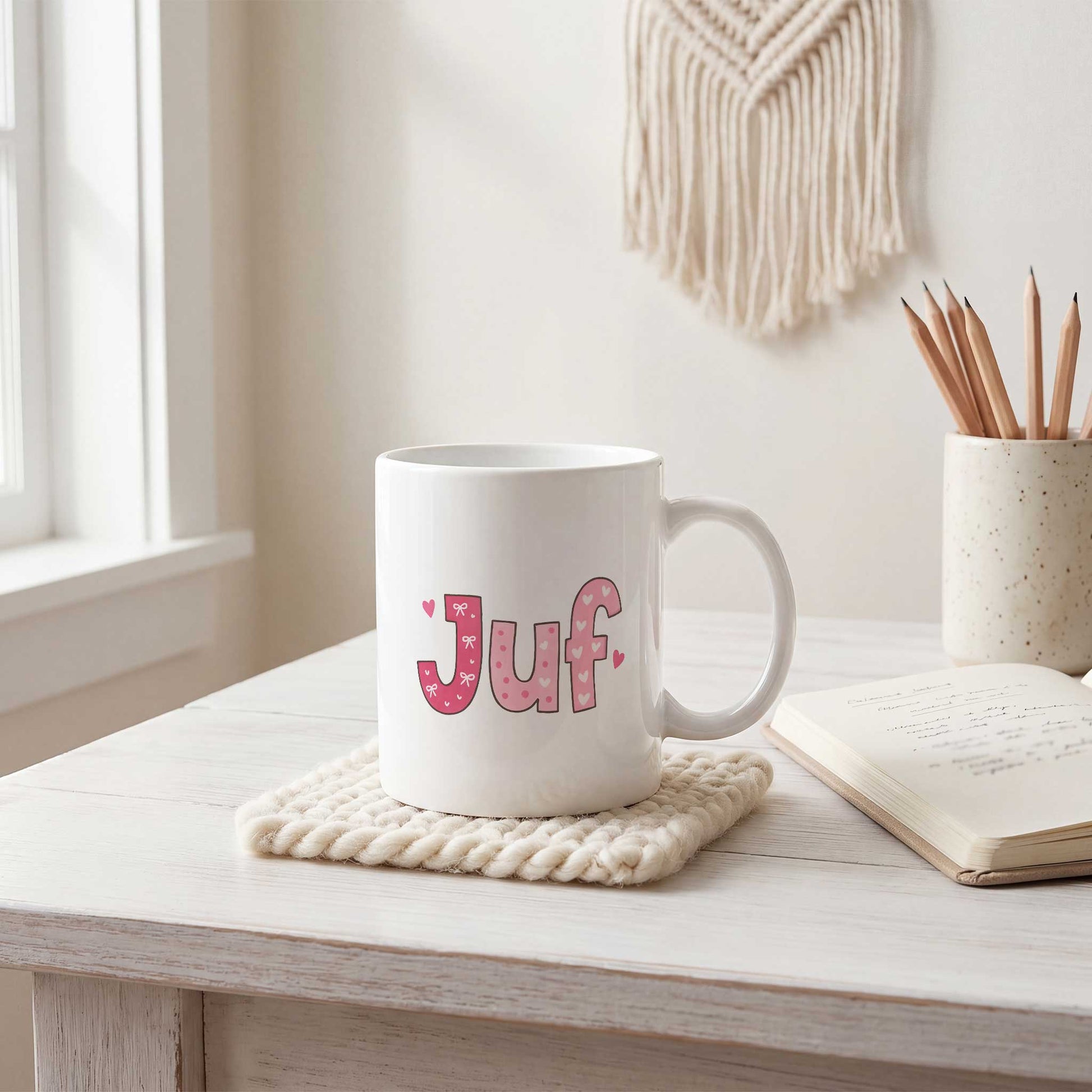Sfeerfoto van een witte mok met roze letters, strikjes en hartjes die samen de tekst Juf vormen op een licht bureau naast een notitieboek en een pot met potloden