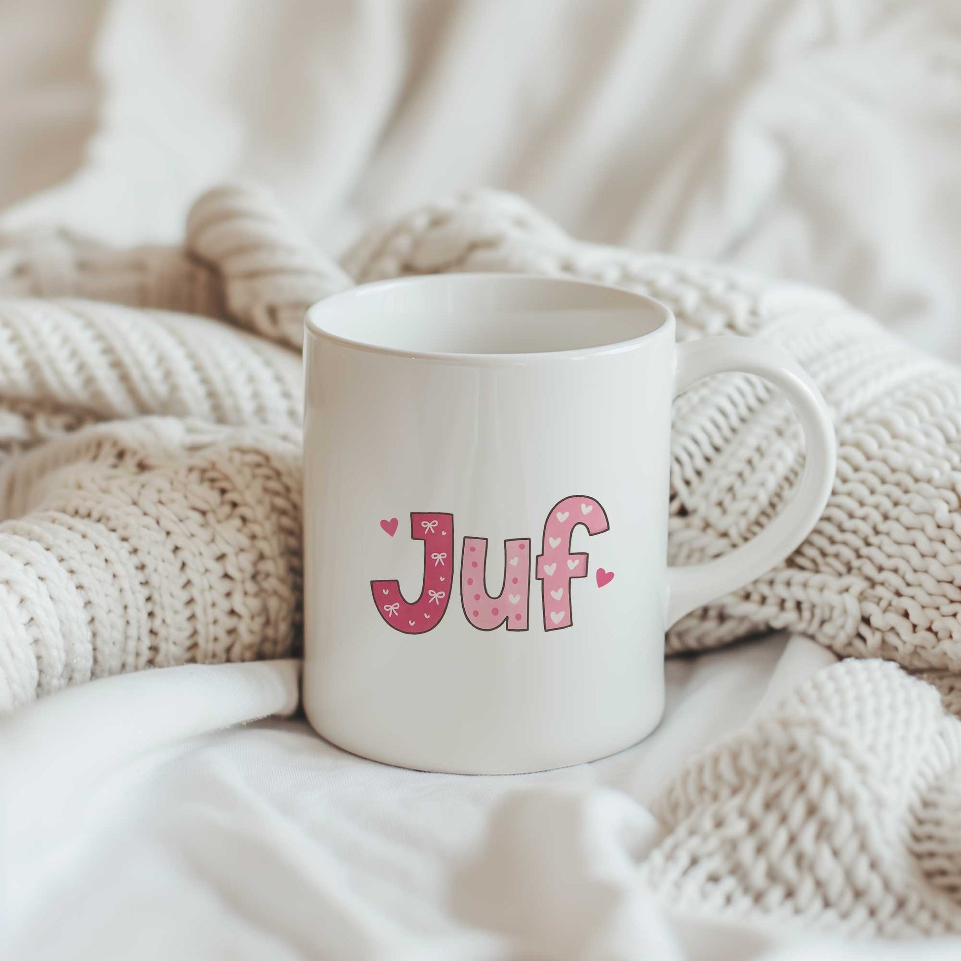 Sfeerfoto van een witte mok met roze letters, strikjes en hartjes die samen de tekst Juf vormen op een lichte gebreide deken