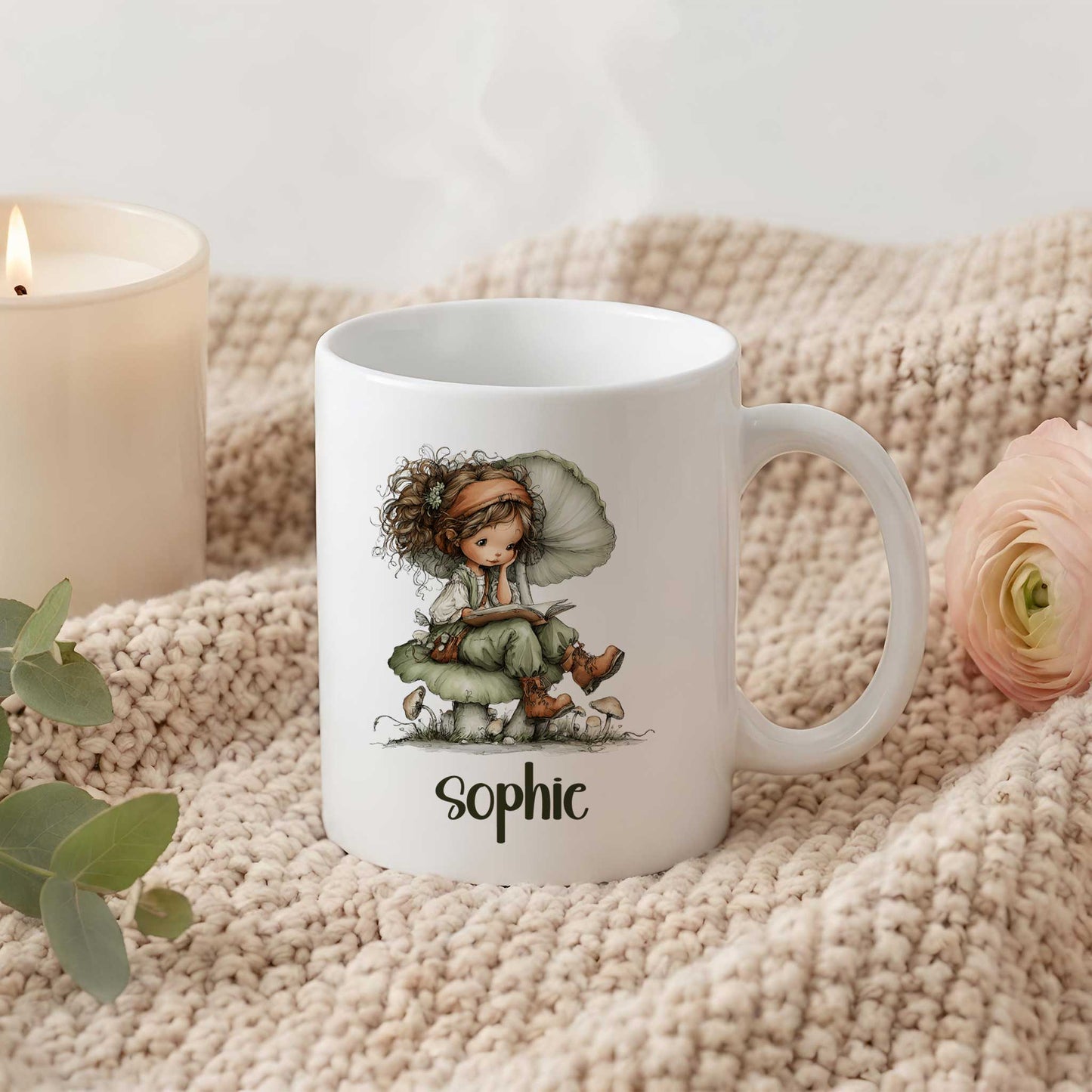 Witte mok met illustratie van een lezende fee op een paddenstoel en de naam Sophie op een gebreide beige deken naast een kaars en bloem.